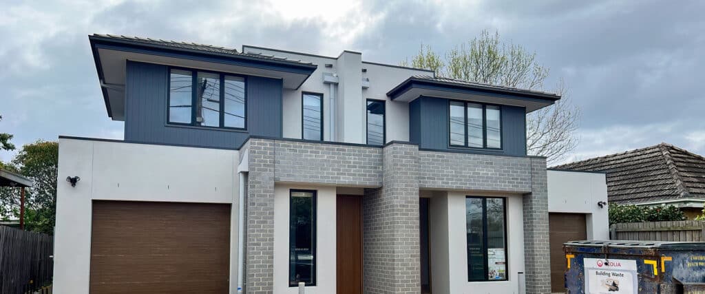 Duplex moderne bicolore: brique grise, bardage bleu sombre et enduit clair. Portes de garage bois. Benne de construction « VEOLIA Building Waste ».