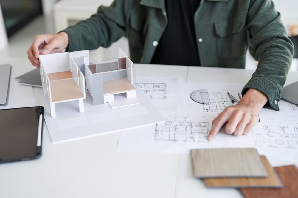 Conception architecturale: travail sur maquette et plans. Designer pointant des plans d'étage détaillés tout en manipulant une maquette architecturale minimaliste avec sols en bois.