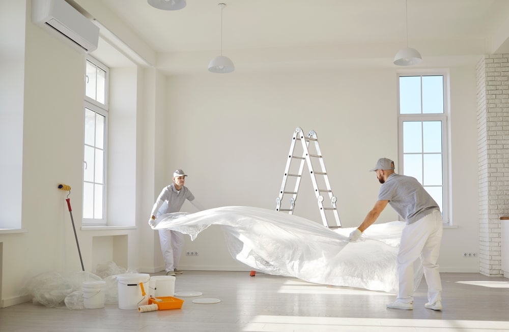 Peinture et rénovation : Préparation professionnelle du sol. Deux ouvriers en tenue de travail déploient une bâche de protection au sol d'une grande salle lumineuse pour la rénovation et la peinture.